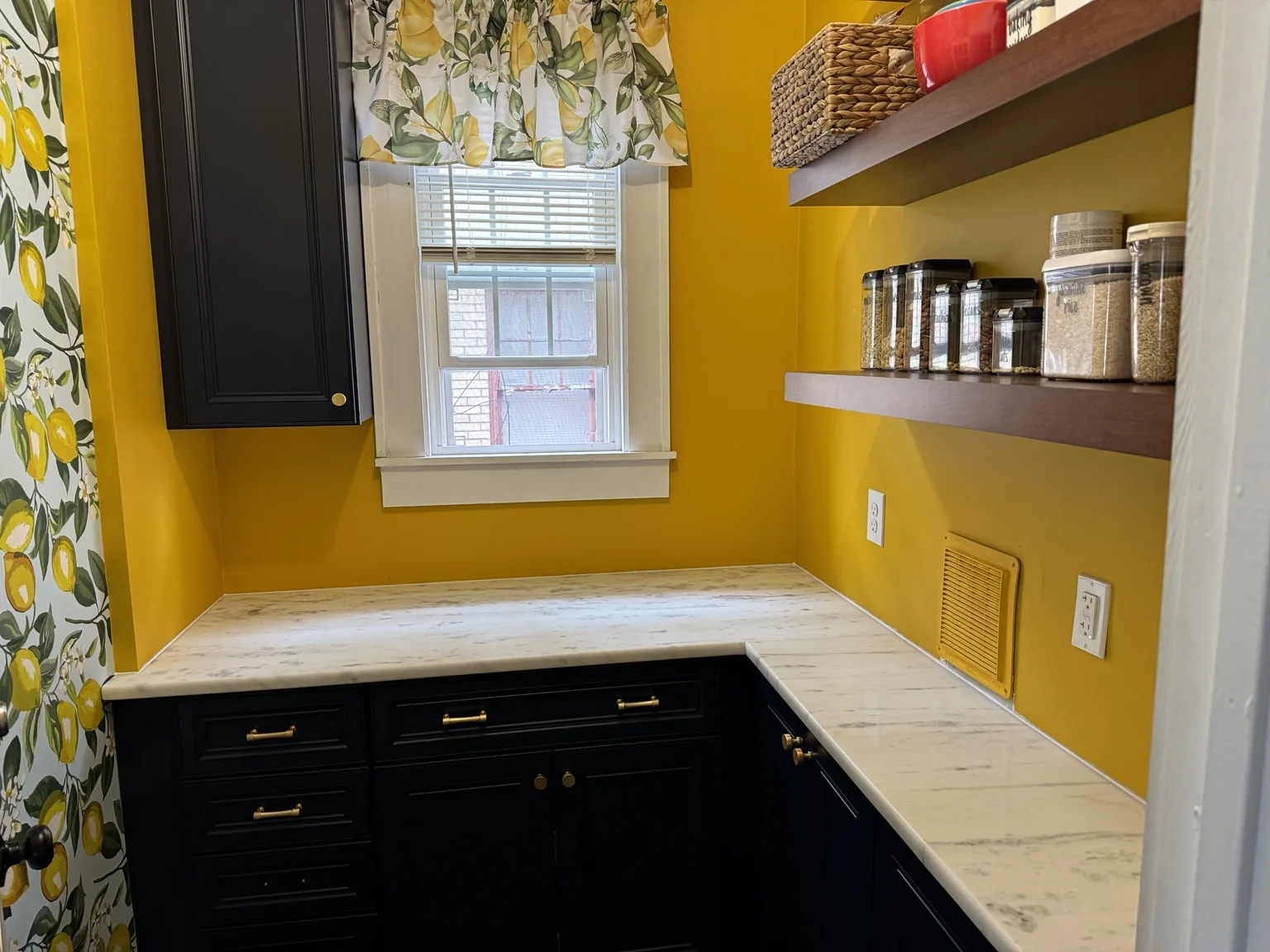 Pantry with finished white stone countertop, dark cabinetry, wood shelves, and lemon window valance