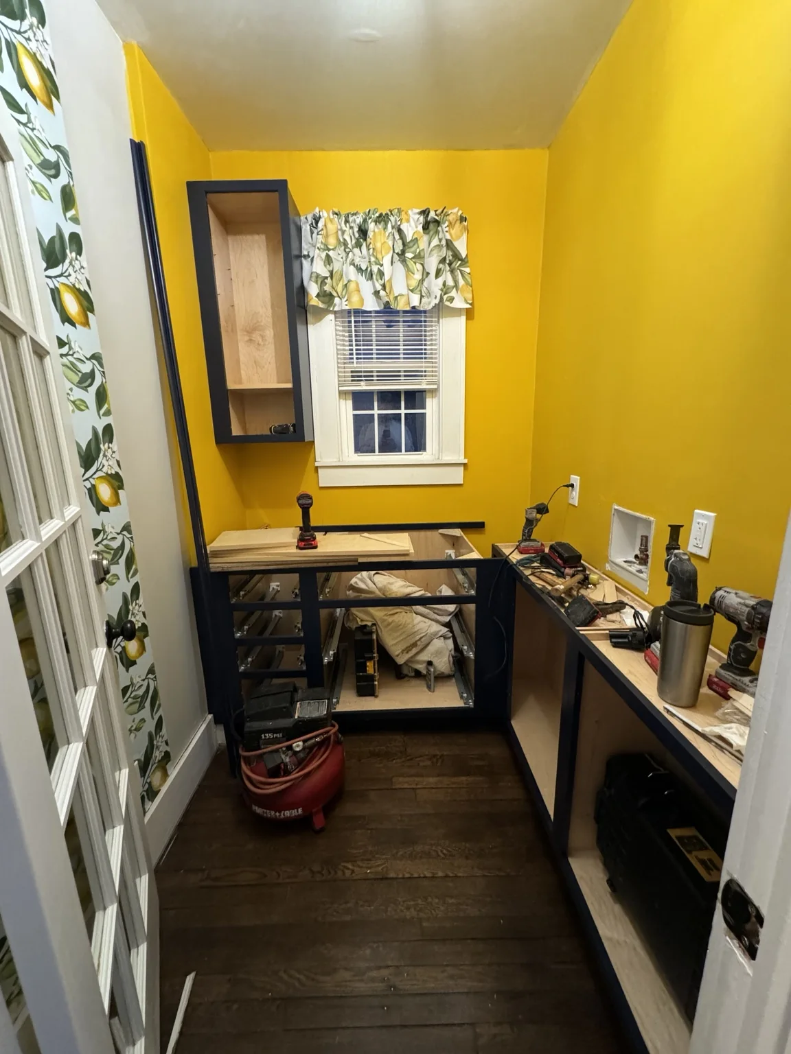 Pantry under construction with dark cabinetry, tools, and lemon-print fabric at the window
